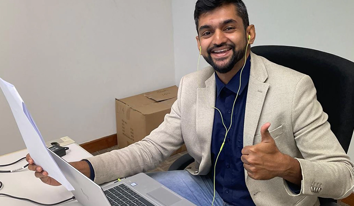 Smiling man in a blazer with earbuds gives thumbs up while sitting at a desk with a laptop and papers.